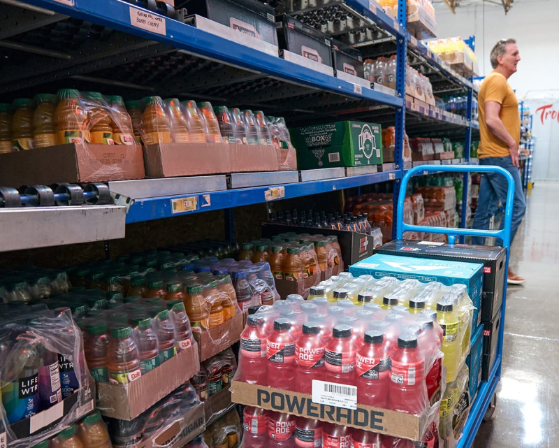 A vending operator warehouse showing racks of beverages with a trolley in the foreground.